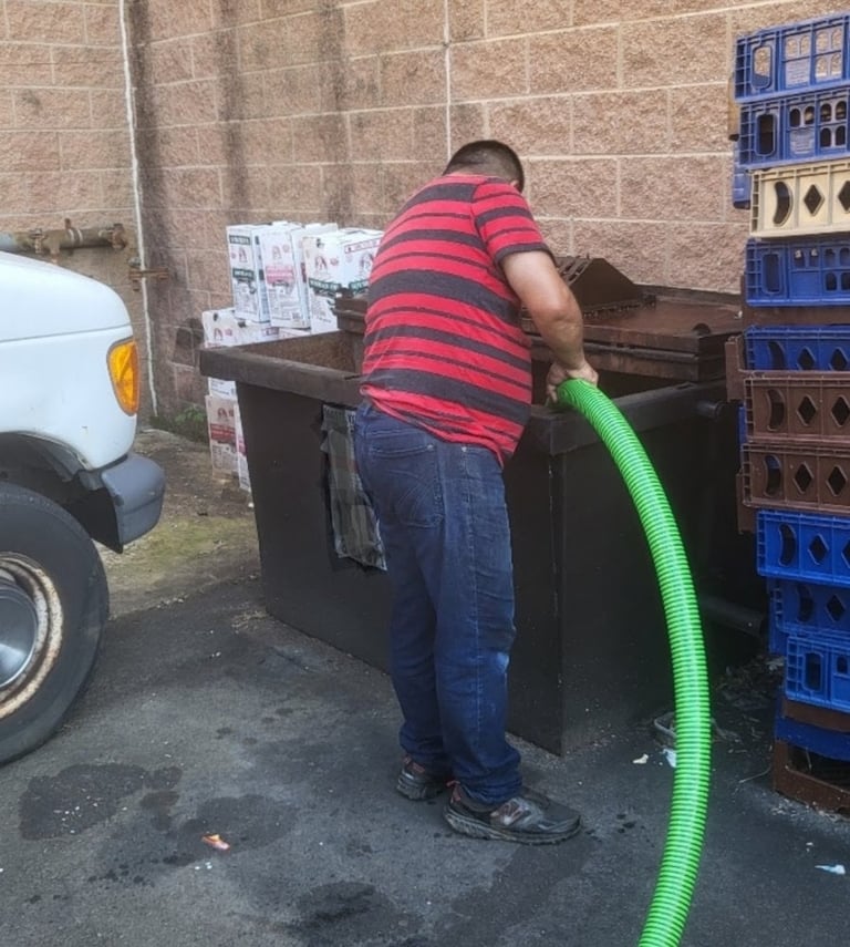 Person in red striped shirt using green hose to fill or clean a dumpster against brick wall