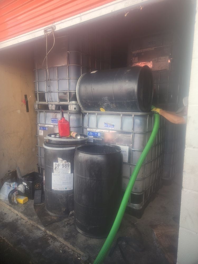 Storage shed containing large black water tanks, metal barrel with red lid, green hose, and various industrial containers under red corrugated roof
