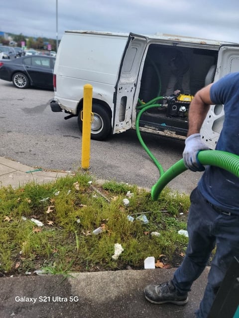 Worker holding green hose connected to white service van with equipment, yellow post visible in foreground